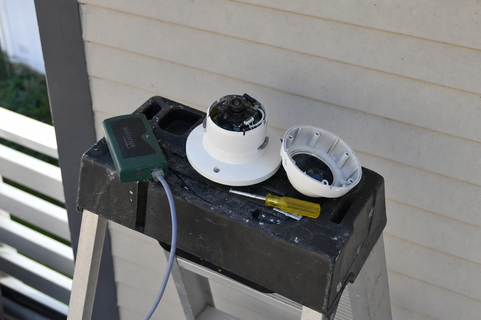 The top tray of a step ladder against a weatherboard wall, holding an opened white dome camera with its cover lying off to one side, a cable tester with a lead plugged into it, and a screwdriver